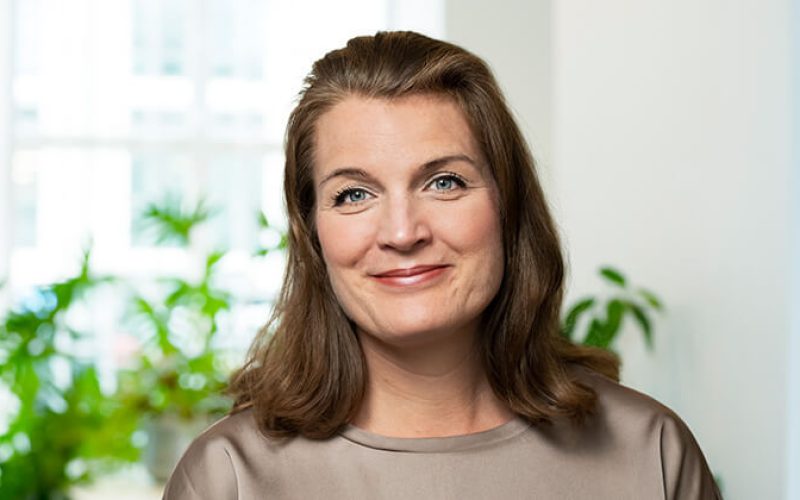 En kvinna med axellångt brunt hår och ljusbrun blus ler mjukt mot kameran. Hon befinner sig inomhus med gröna växter och ett ljust fönster i bakgrunden.