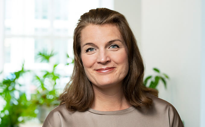 En kvinna med axellångt brunt hår och ljusbrun blus ler mjukt mot kameran. Hon befinner sig inomhus med gröna växter och ett ljust fönster i bakgrunden.