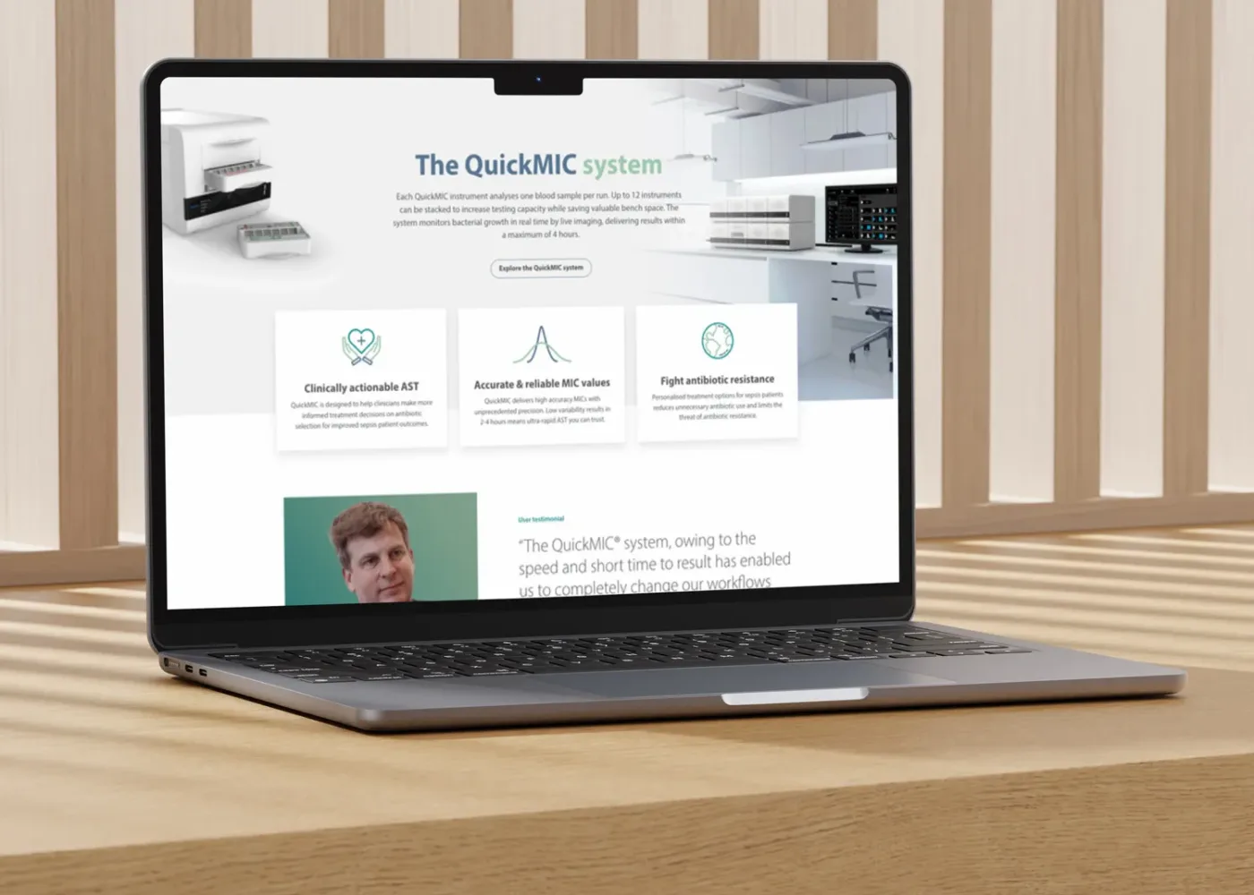 En bärbar dator på ett träbord visar en webbplats som marknadsför QuickMIC-systemet, med text om kliniska fördelar, ikoner och ett vittnesmål med ett foto av en man. Ljusa träribbor bildar bakgrunden.