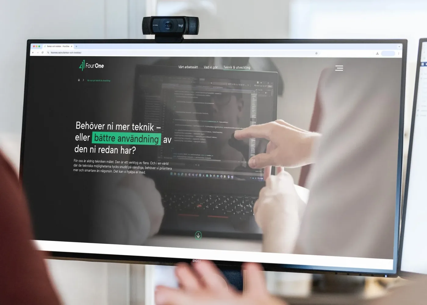 Två personer sitter framför en datorskärm som visar en hemsida med svensk text och kod. En person pekar på skärmen och en kaffemugg syns på skrivbordet. En webbkamera är monterad ovanpå bildskärmen.