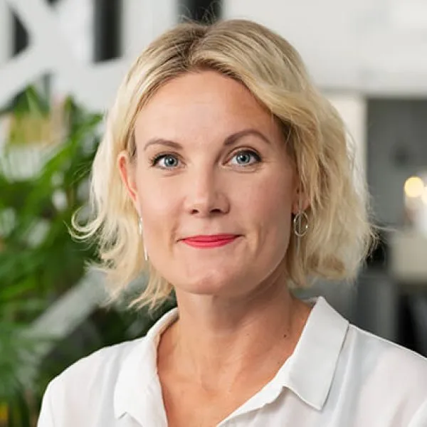 En kvinna med kort blont hår, vit blus och ringörhängen ler mjukt när hon står inomhus med gröna växter och suddig modern inredning i bakgrunden.