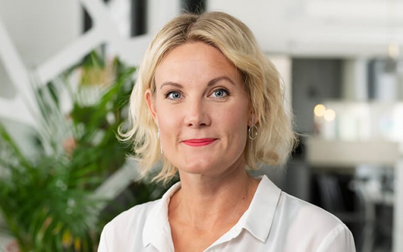 En kvinna med kort blont hår, vit blus och ringörhängen ler mjukt när hon står inomhus med gröna växter och suddig modern inredning i bakgrunden.