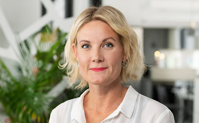 En kvinna med kort blont hår, vit blus och ringörhängen ler mjukt när hon står inomhus med gröna växter och suddig modern inredning i bakgrunden.