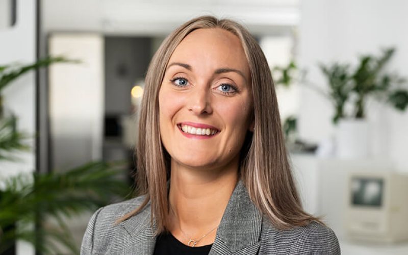 En leende kvinna med rakt, ljusbrunt hår och grå kavaj står i ett ljust, modernt kontor med växter och vit inredning i bakgrunden.