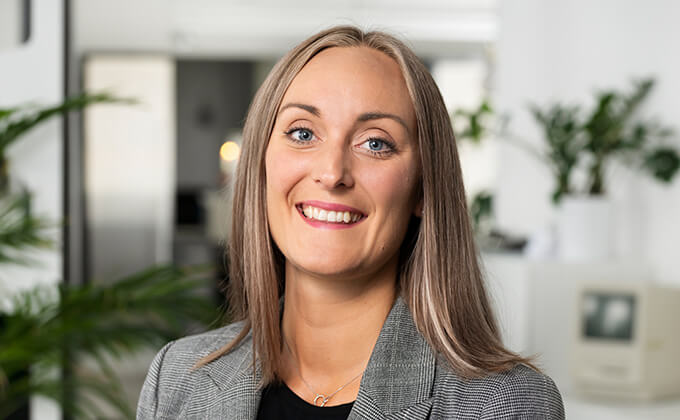 En leende kvinna med rakt, ljusbrunt hår och grå kavaj står i ett ljust, modernt kontor med växter och vit inredning i bakgrunden.