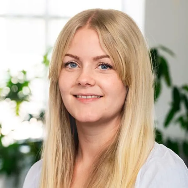 En ung kvinna med långt blont hår och ljus skjorta ler mot kameran. Bakgrunden är mjukt upplyst och visar gröna växter och ett ljust fönster.