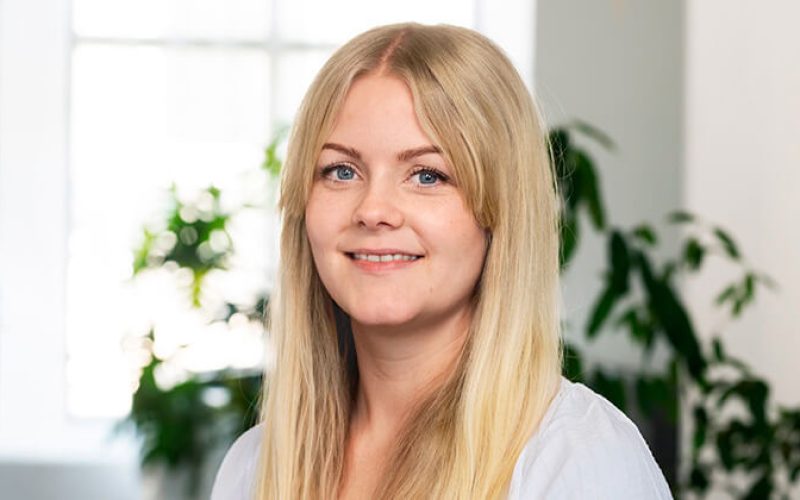 En ung kvinna med långt blont hår och ljus skjorta ler mot kameran. Bakgrunden är mjukt upplyst och visar gröna växter och ett ljust fönster.