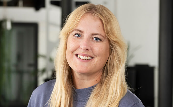 Leende kvinna med långt blont hår och grå topp står inomhus mot en suddig bakgrund med fönster och växter.