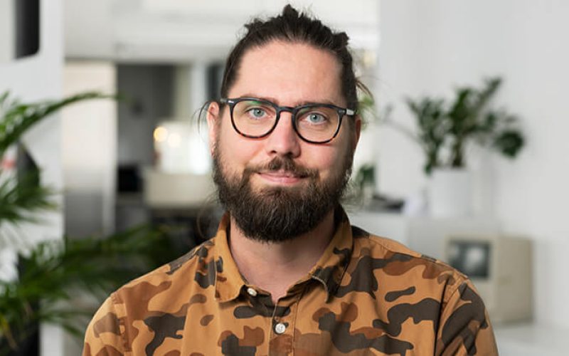 En man med glasögon, skägg och långt bakåtkammat hår, klädd i en brun kamouflageskjorta, står i ett starkt upplyst rum med växter och en suddig bakgrund.