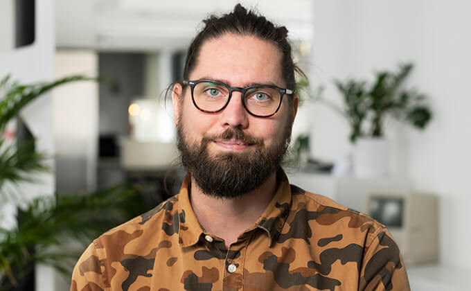 En man med glasögon, skägg och långt bakåtkammat hår, klädd i en brun kamouflageskjorta, står i ett starkt upplyst rum med växter och en suddig bakgrund.