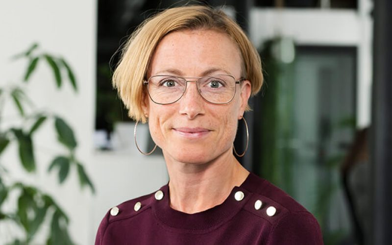 En kvinna med kort blont hår, stora runda glasögon, örhängen och en rödbrun topp med dekorativa knappar på axlarna ler mot kameran. Bakom henne syns en grön växt och en suddig kontorsbakgrund.