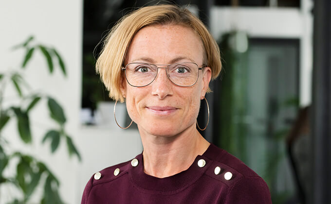En kvinna med kort blont hår, stora runda glasögon, örhängen och en rödbrun topp med dekorativa knappar på axlarna ler mot kameran. Bakom henne syns en grön växt och en suddig kontorsbakgrund.