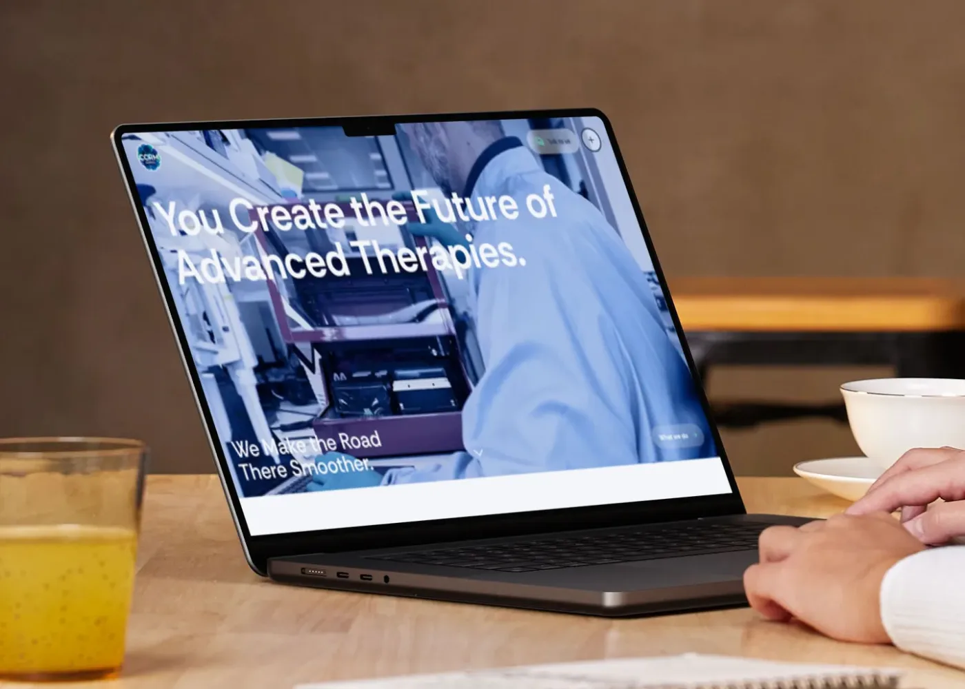 En person använder en bärbar dator vid ett träbord med en anteckningsbok, penna, ett glas juice och en skål. Den bärbara datorns skärm visar en webbplats med texten 