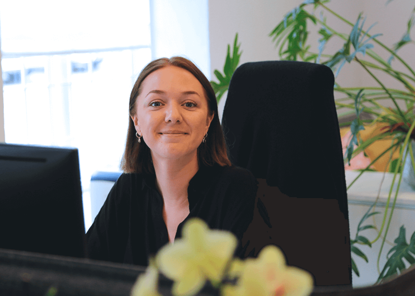 En kvinna med kort brunt hår, klädd i en svart topp, sitter vid ett skrivbord på ett kontor och ler mot kameran. I bakgrunden syns gröna växter och en datorskärm.