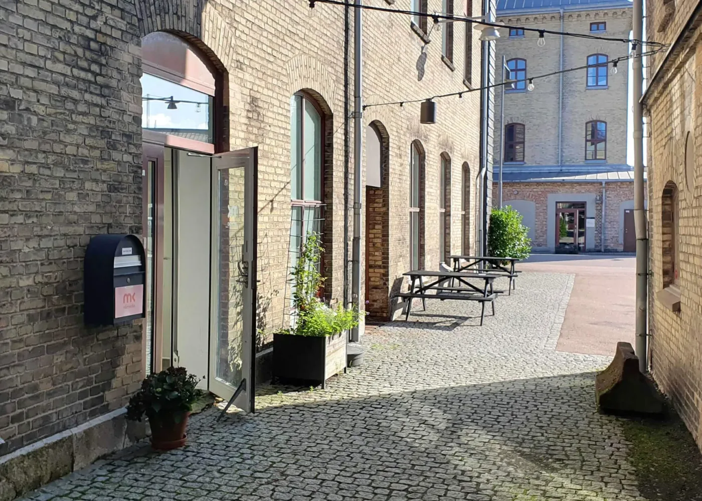 En solbelyst kullerstensgränd med tegelbyggnader, en öppen glasdörr, krukväxter, ljusslingor och picknickbord, som leder till en innergård och en annan byggnad i bakgrunden.