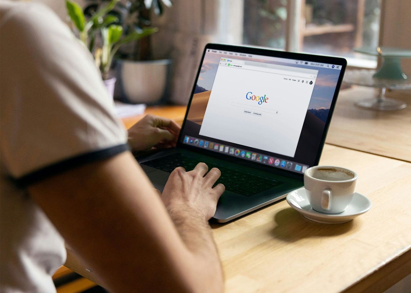 En person skriver på en bärbar dator som visar Googles sökstartsida, sittande vid ett träskrivbord med en kopp kaffe, ett anteckningsblock och krukväxter i närheten.