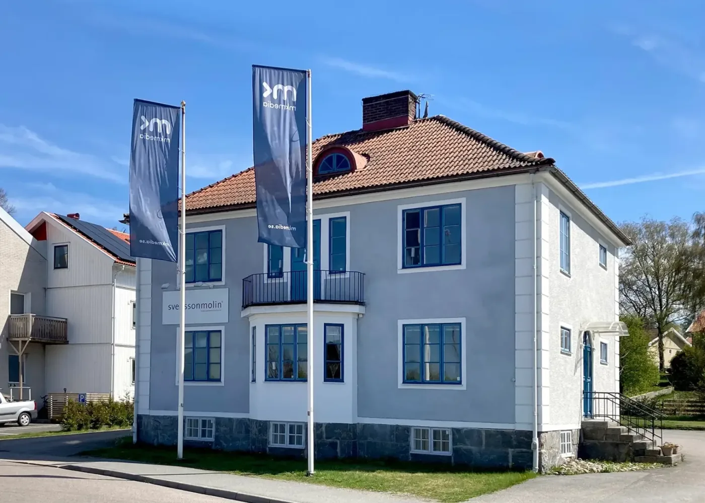 En ljusblå tvåvåningsbyggnad med rött tegeltak, blå fönsterkarmar och två höga flaggor utanför. Byggnaden ligger på en hörntomt under en klarblå himmel.