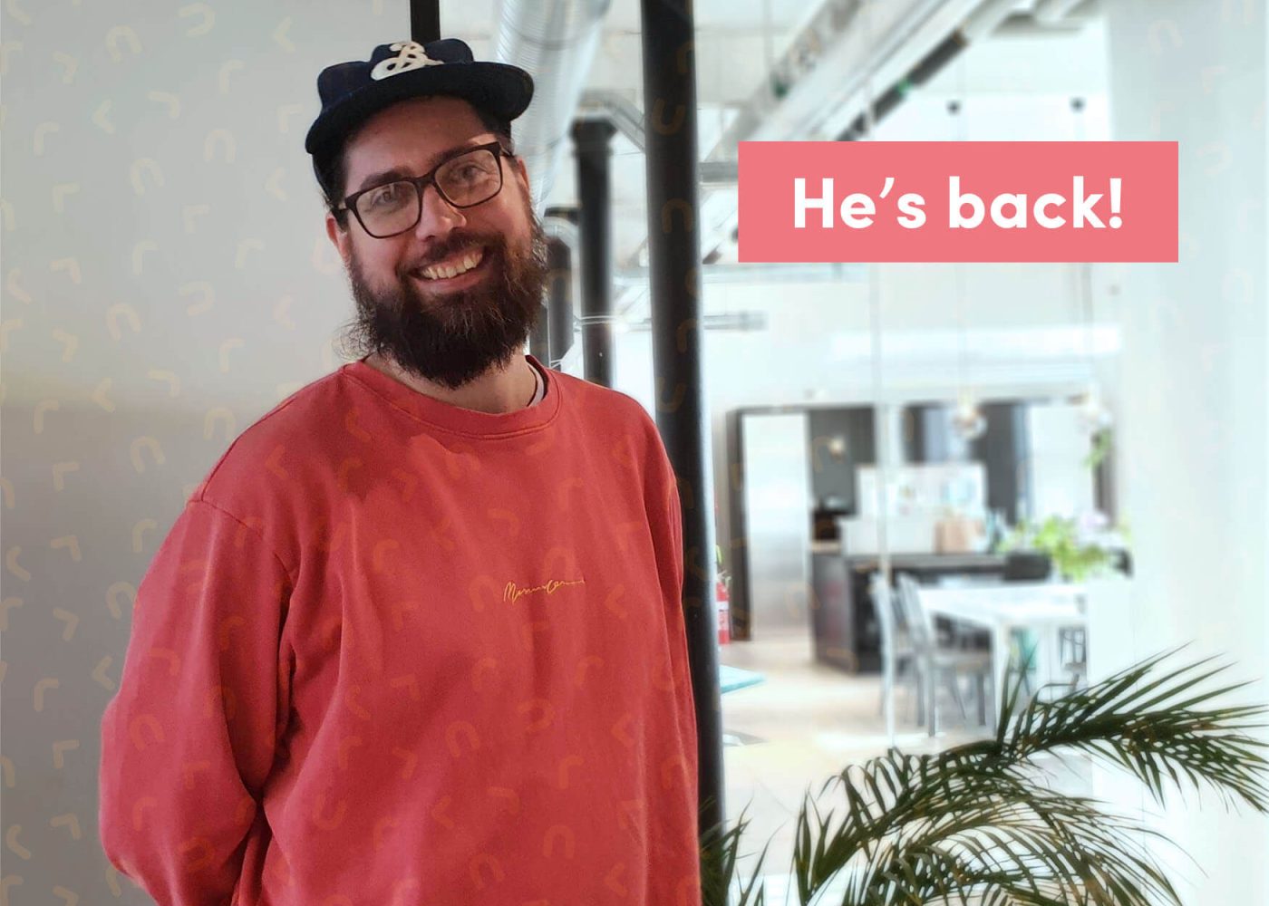 En leende skäggig man med glasögon och keps står i en modern, ljus kontorslokal. Han bär en röd sweatshirt. Rosa text på bilden lyder Hes back! En grön växt syns i förgrunden.