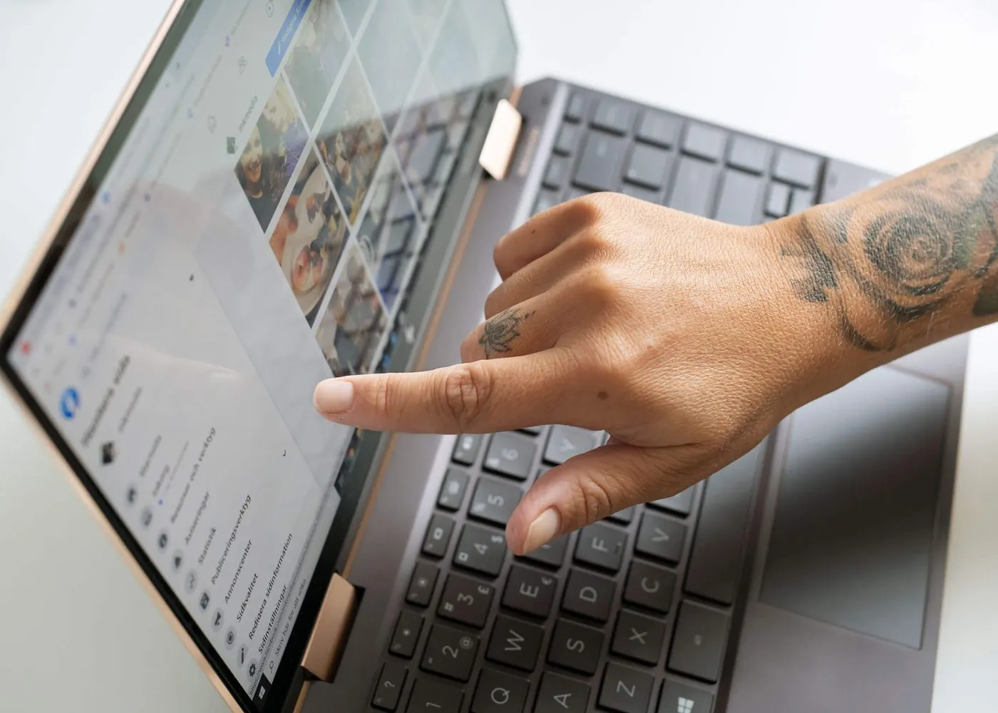 En tatuerad hand pekar på en laptopskärm som visar ett fotogalleri och menyalternativ, med en del av ett svart tangentbord och enhetens roséguldfärgade gångjärn synliga.