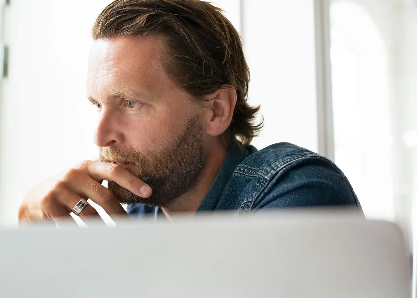 En man med skägg och långt hår, klädd i jeansskjorta, sitter vid ett skrivbord och tittar eftertänksamt på en laptopskärm med handen vilande på hakan. Bakgrunden är fylld av starkt, naturligt ljus.