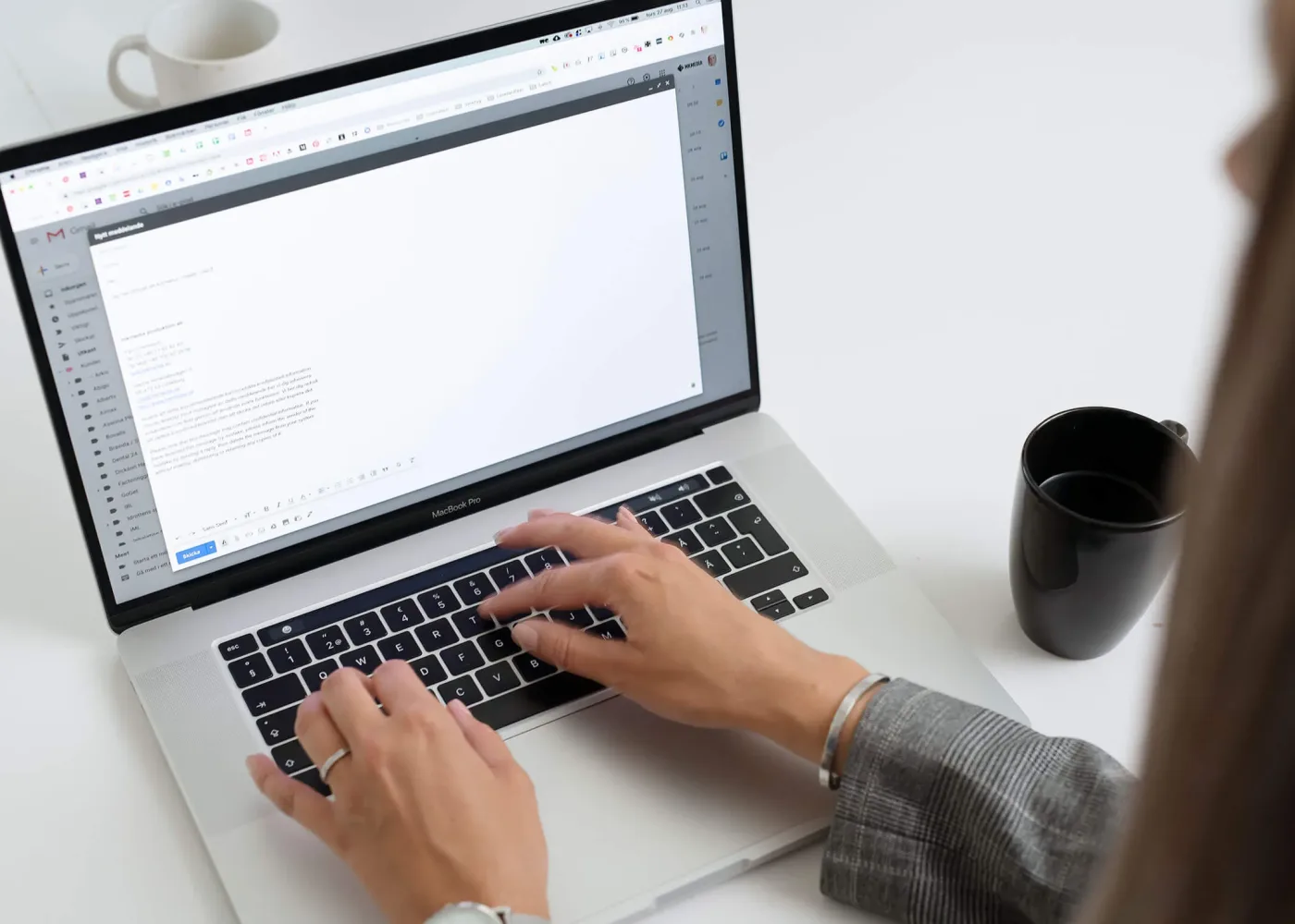 En person skriver ett e-postmeddelande på en bärbar dator vid ett skrivbord med en svart kaffemugg i närheten. På skärmen visas ett utkast till ett meddelande i Gmail.