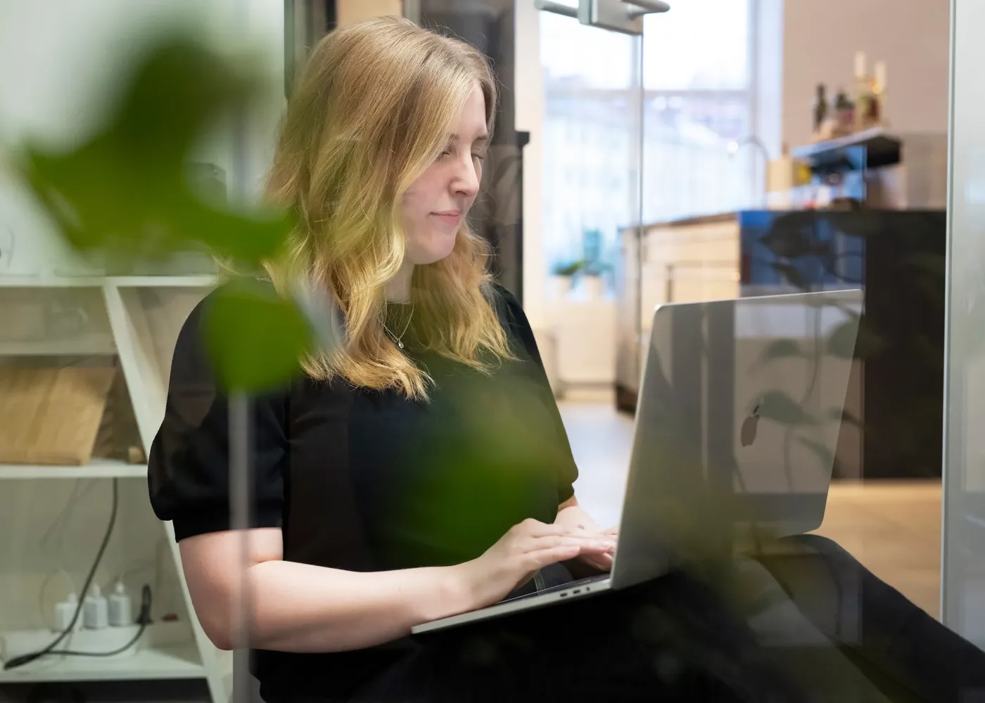 En kvinna med blont hår sitter på ett modernt kontor och arbetar på en laptop. Hon bär en svart topp och ser fokuserad ut. I förgrunden står några gröna växter och i bakgrunden finns hyllor och ett kök.