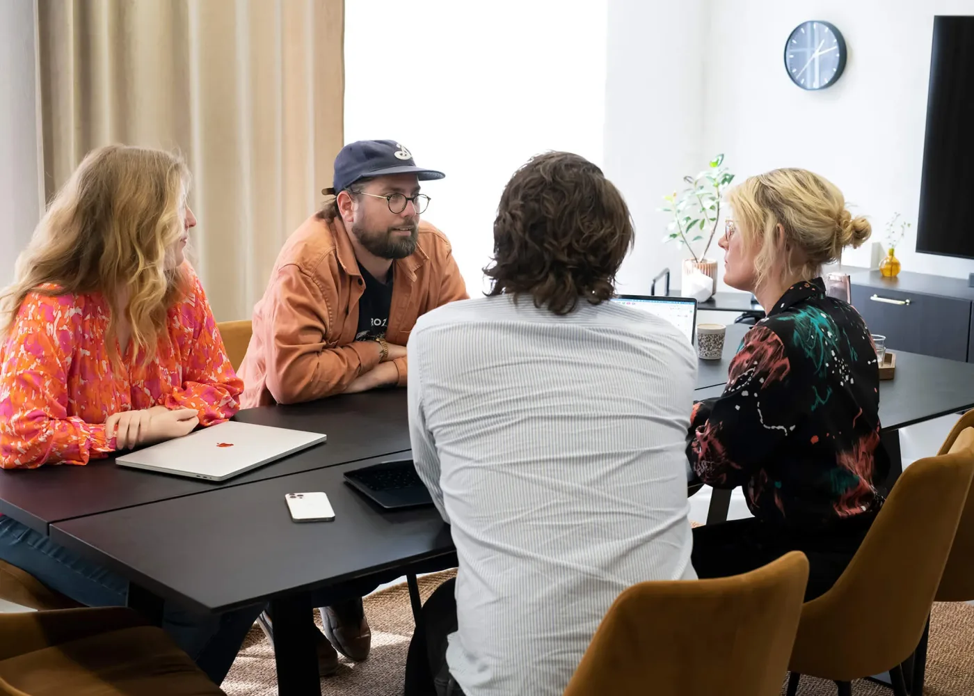 Fyra personer sitter runt ett bord på ett kontor och diskuterar. Två bärbara datorer och en smartphone står på bordet, och på väggen i bakgrunden hänger en klocka och en TV.