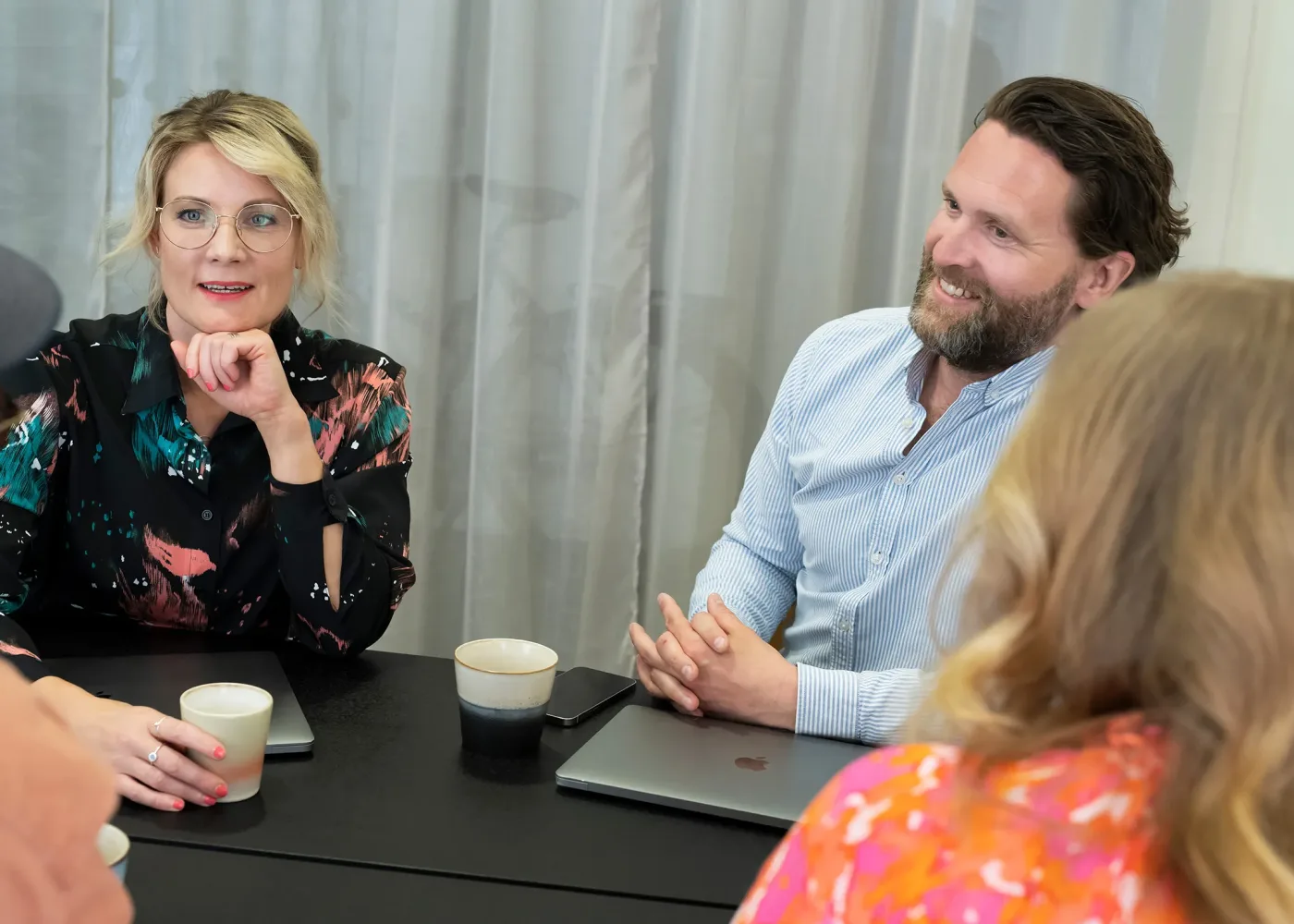 Fyra personer sitter runt ett bord och diskuterar. Två av dem har bärbara datorer framför sig och kaffekoppar står på bordet. De verkar engagerade och ler. En skir gardin hänger i bakgrunden.