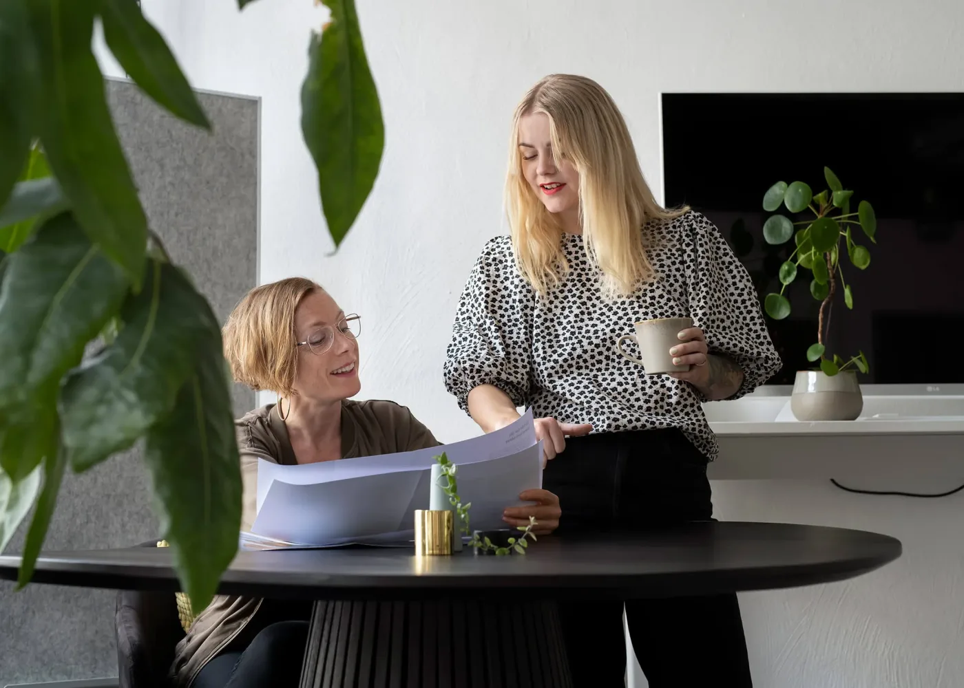 Två kvinnor i en kontorsmiljö granskar dokument tillsammans vid ett runt bord, där den ena sitter och den andra står och håller i en mugg. Gröna växter syns i förgrunden och bakgrunden.