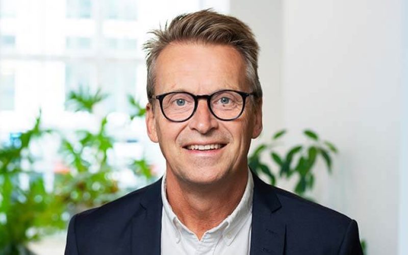 En leende medelålders man med glasögon och kort, ljusbrunt hår, klädd i mörk kavaj och vit skjorta, står inomhus med gröna växter och stora fönster i bakgrunden.