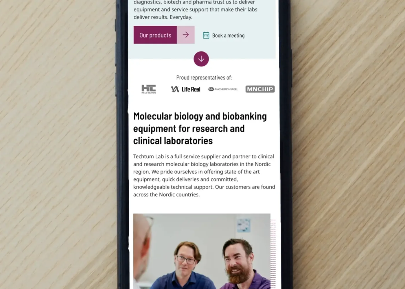 En smartphone på en yta av ljust trä visar en webbplats om molekylärbiologi och biobanksutrustning, med text och ett foto av två personer i samtal längst ner på skärmen.