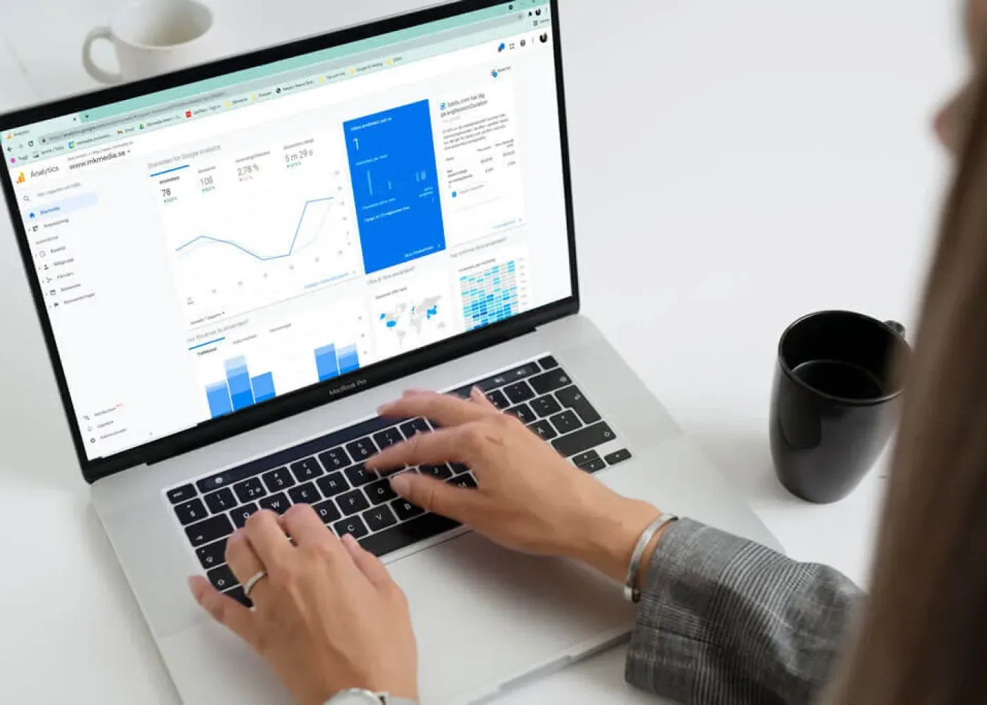 En person skriver på en bärbar dator som visar analytiska instrumentpaneler med grafer och diagram. En svart kaffemugg och en vit kopp står på det vita skrivbordet bredvid den bärbara datorn.